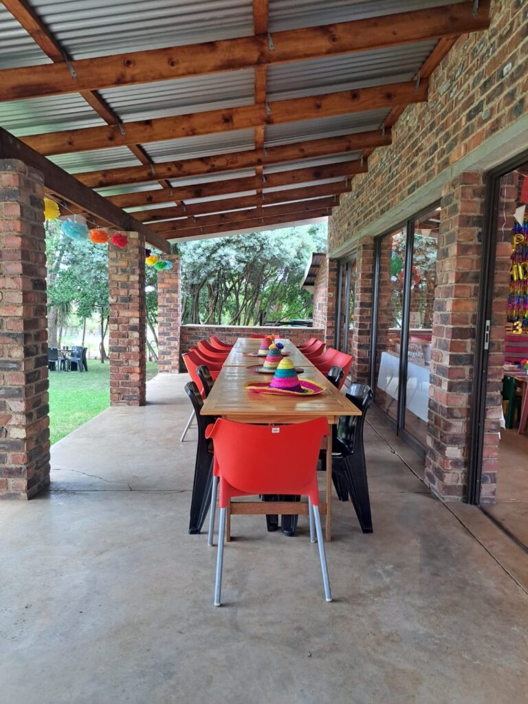 A festive setup at The Barn on Boundary Party Venue with a long wooden table, colorful chairs, and decorations hanging from the ceiling.