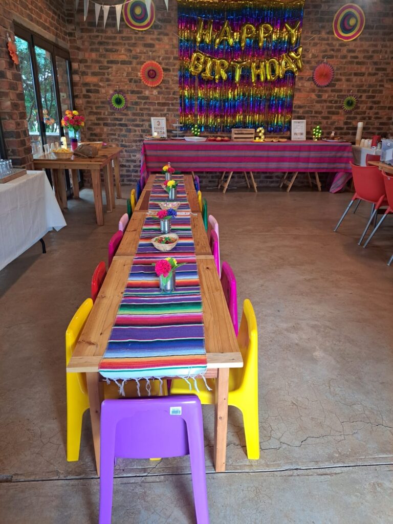A colorful birthday party setup at The Barn on Boundary in Johannesburg, Northriding, featuring a long wooden table with multicolored chairs and vibrant decorations.
