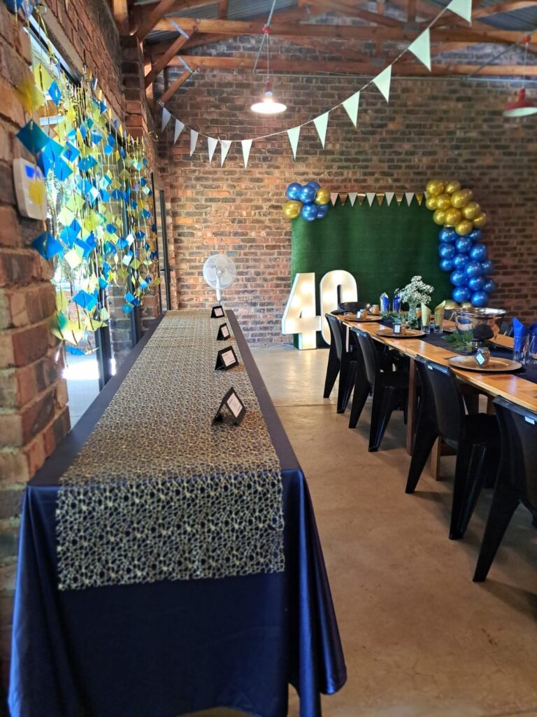 A decorated party venue at the Barn on Boundary in Johannesburg, Northriding, featuring a long table with a leopard print runner, blue tablecloth, and seating arrangements. The brick walls are adorned with colorful decorations and β40β sign.