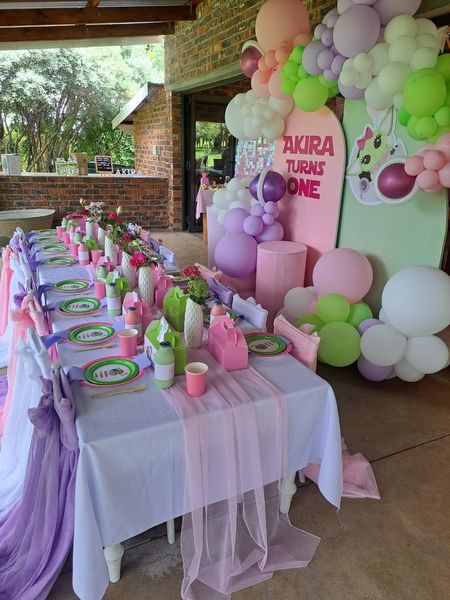 A colorful setup for a first birthday party at the Barn on Boundary Party Venue in Johannesburg, Northriding, featuring a long table adorned with pink and green decorations and a backdrop of balloons beside a banner celebrating Akiraβs first birthday.