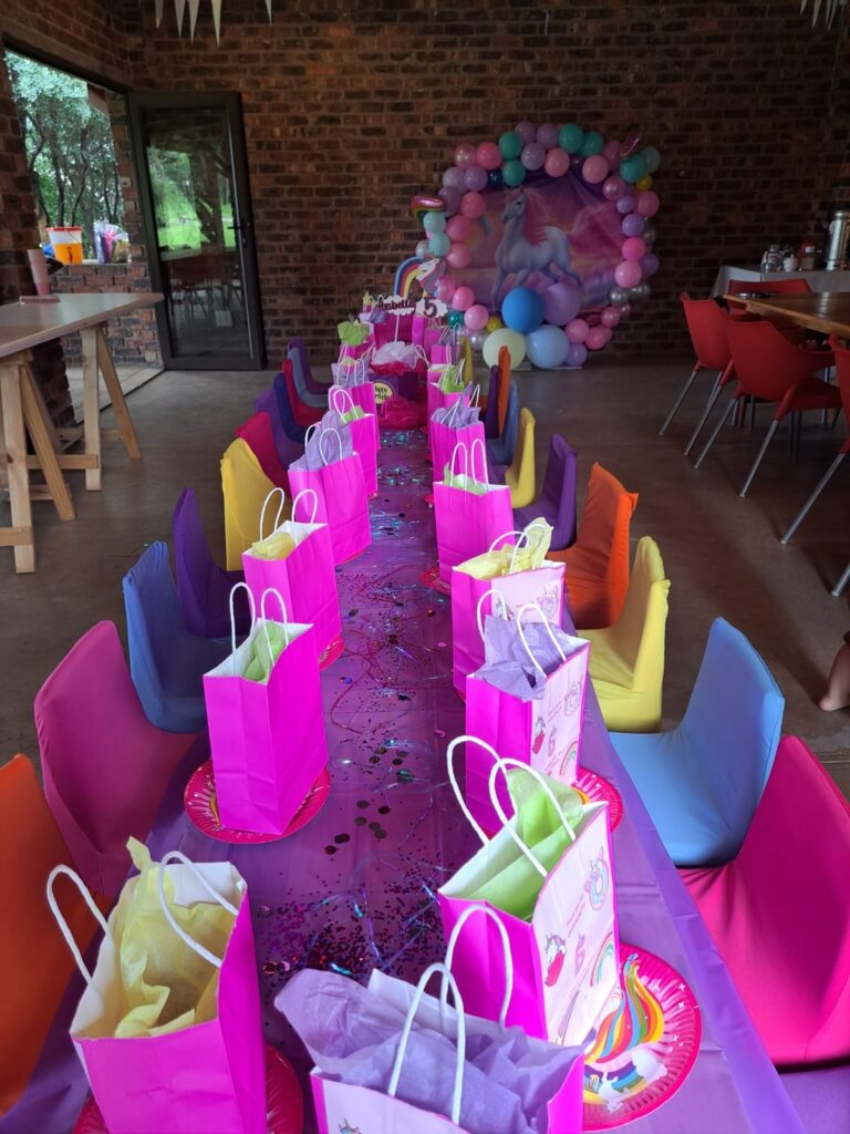 A colorful party setup at The Barn on Boundary in Johannesburg, featuring a long table adorned with vibrant gift bags and a unicorn-themed backdrop.
