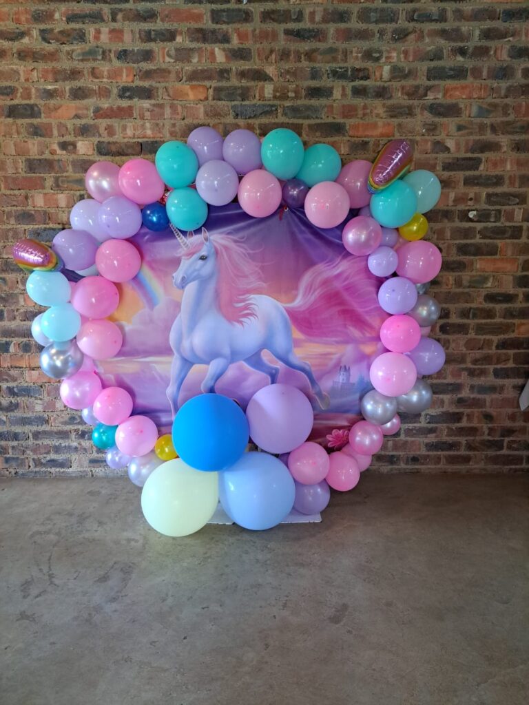 A colorful Balloon Arch unicorn-themed party decoration featuring a mix of pink, purple, and blue balloons arranged around a vibrant unicorn illustration, set against a brick wall at the Barn on Boundary Party Venue in Johannesburg, Northriding.