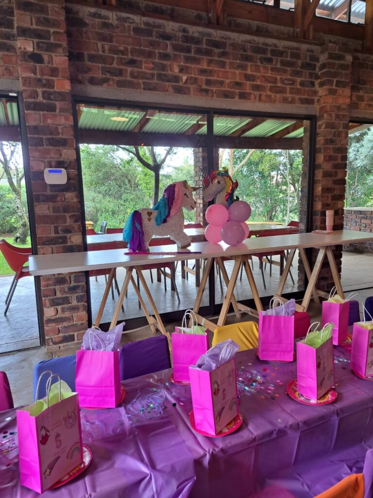 A colorful childrenβs party setup at the Barn on Boundary Party Venue featuring a unicorn piΓ±ata, pink gift bags, and a purple tablecloth.