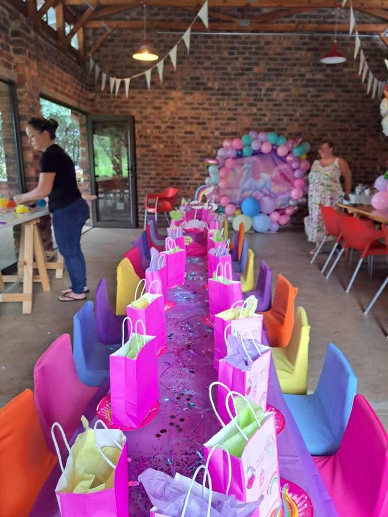 A colorful childrenβs party setup at the Barn on Boundary Party Venue with pink gift bags, a unicorn-themed backdrop, and people preparing.